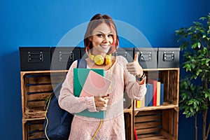 Young caucasian woman wearing student backpack and books smiling happy and positive, thumb up doing excellent and approval sign