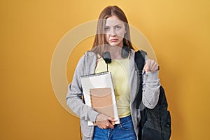 Young caucasian woman wearing student backpack and holding books pointing down looking sad and upset, indicating direction with