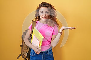 Young caucasian woman wearing student backpack and holding books smiling cheerful presenting and pointing with palm of hand