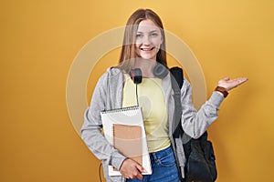 Young caucasian woman wearing student backpack and holding books smiling cheerful presenting and pointing with palm of hand