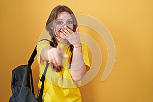 Young caucasian woman wearing student backpack over yellow background laughing at you, pointing finger to the camera with hand