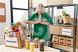 Young caucasian woman wearing volunteer t shirt at donations stand begging and praying with hands together with hope expression on