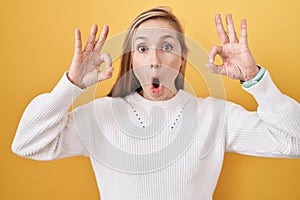 Young caucasian woman wearing white sweater over yellow background looking surprised and shocked doing ok approval symbol with