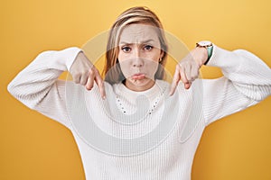 Young caucasian woman wearing white sweater over yellow background pointing down looking sad and upset, indicating direction with