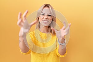 Young caucasian woman wearing yellow sweater shouting frustrated with rage, hands trying to strangle, yelling mad