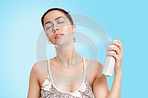 A young Caucasian woman with wet skin splashes thermal water on her face, closing her eyes from the drops. Blue background. The