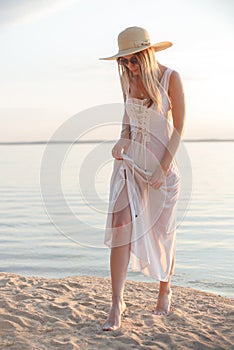 Young caucasian woman in white dress, summer hat and sunglasses posing on beach on sunset