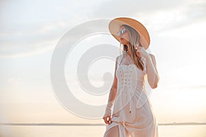 Young caucasian woman in white dress, summer hat and sunglasses posing on beach on sunset