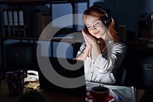 Young caucasian woman working at the office at night sleeping tired dreaming and posing with hands together while smiling with