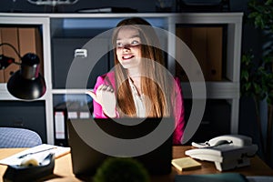 Young caucasian woman working at the office at night smiling with happy face looking and pointing to the side with thumb up
