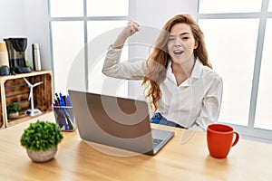 Young caucasian woman working at the office using computer laptop angry and mad raising fist frustrated and furious while shouting
