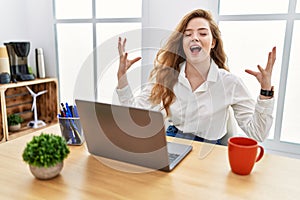 Young caucasian woman working at the office using computer laptop celebrating mad and crazy for success with arms raised and