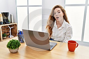 Young caucasian woman working at the office using computer laptop in shock face, looking skeptical and sarcastic, surprised with
