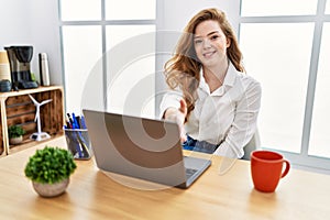 Young caucasian woman working at the office using computer laptop smiling friendly offering handshake as greeting and welcoming