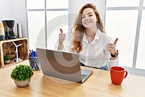 Young caucasian woman working at the office using computer laptop success sign doing positive gesture with hand, thumbs up smiling