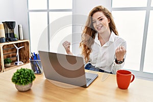 Young caucasian woman working at the office using computer laptop very happy and excited doing winner gesture with arms raised,