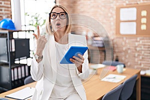 Young caucasian woman working at the office wearing glasses amazed and surprised looking up and pointing with fingers and raised