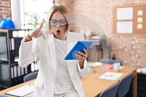 Young caucasian woman working at the office wearing glasses pointing down with fingers showing advertisement, surprised face and