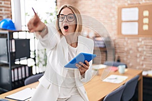 Young caucasian woman working at the office wearing glasses pointing with finger surprised ahead, open mouth amazed expression,