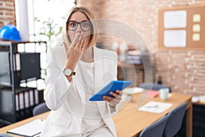 Young caucasian woman working at the office wearing glasses shocked covering mouth with hands for mistake