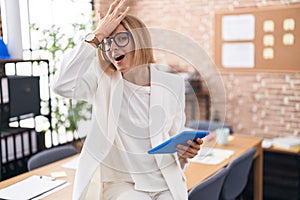 Young caucasian woman working at the office wearing glasses surprised with hand on head for mistake, remember error