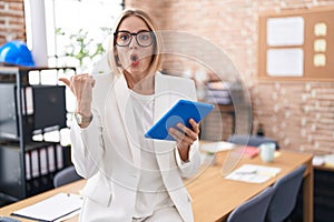 Young caucasian woman working at the office wearing glasses surprised pointing with hand finger to the side, open mouth amazed