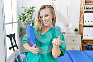 Young caucasian woman working at pain recovery clinic beckoning come here gesture with hand inviting welcoming happy and smiling