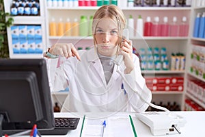 Young caucasian woman working at pharmacy drugstore speaking on the telephone pointing down looking sad and upset, indicating