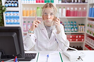 Young caucasian woman working at pharmacy drugstore speaking on the telephone pointing up looking sad and upset, indicating