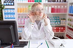 Young caucasian woman working at pharmacy drugstore speaking on the telephone smiling with hand over ear listening an hearing to