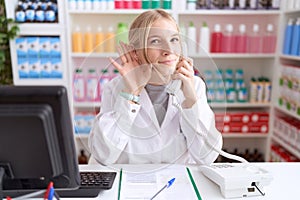 Young caucasian woman working at pharmacy drugstore speaking on the telephone smiling with hand over ear listening an hearing to