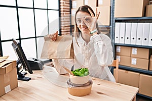 Young caucasian woman working at small business ecommerce holding take away food smiling happy doing ok sign with hand on eye