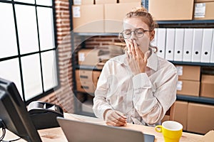 Young caucasian woman working at small business ecommerce using laptop bored yawning tired covering mouth with hand