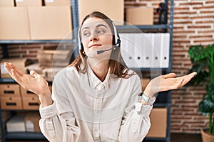 Young caucasian woman working at small business ecommerce wearing headset clueless and confused expression with arms and hands