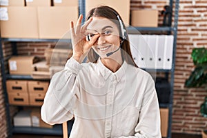 Young caucasian woman working at small business ecommerce wearing headset smiling happy doing ok sign with hand on eye looking
