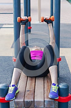 Young caucasian woman workouts on the park sports ground with dumbbells, laying on the sport bar