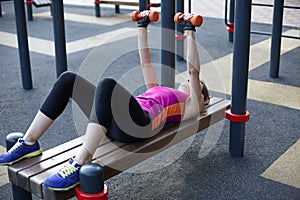 Young caucasian woman workouts on the park sports ground with dumbbells, laying on the sport bar