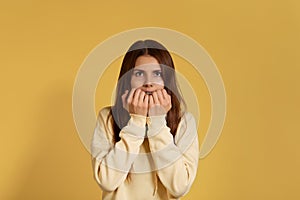 Young caucasian woman in a yellow hoodie holds hands on chin, surprised to hear news, shocked, dissatisfied, amazed by sudden news