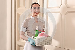 Young chambermaid holding basket with cleaning products in hotel. Space for text