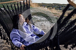 A young charming Asian woman lies in a hammock and looks at the mountains. A beautiful Korean woman in casual clothes enjoys