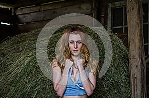 Young charming blonde girl with long hair in a barn on a farm near a haystack in the countryside with alluring eyes is wearing a