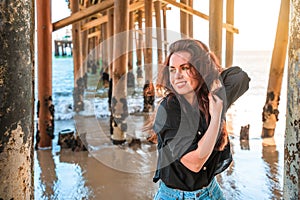 A young charming brunette woman in a black shirt and denim shorts poses under a pier on the beach of Malibu, California