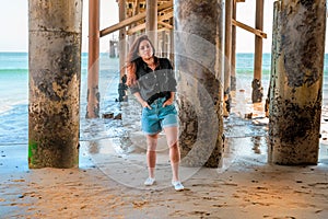 A young charming brunette woman in a black shirt and denim shorts poses under a pier on the beach of Malibu, California