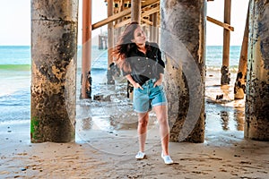 A young charming brunette woman in a black shirt and denim shorts poses under a pier on the beach of Malibu, California