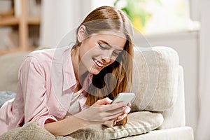 Young charming girl, lying on sofa, smiling writing sms on cell phone.