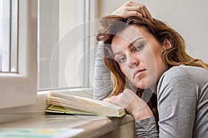 Young charming girl student, with long hair, sad at the window reading book with lessons