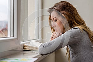 Young charming girl student, with long hair, sad at the window reading book with lessons