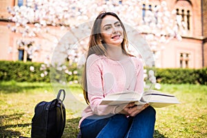 Young, charming girl student reads book against background of green grass and blossom tree