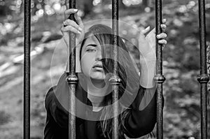 Young charming girl the teenager with long hair sitting behind bars in prison prisoner in a medieval jail with sad, pleading eyes