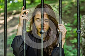 Young charming girl the teenager with long hair sitting behind bars in prison prisoner in a medieval jail with sad, pleading eyes